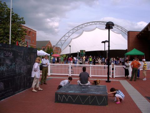 1971 Ford Ranchero - A crowd gathers on a brick plaza in front of a white tensile-fabric amphitheater pavilion under cloudy skies, with white fencing, vendor ca…