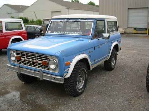 1975 Ford Bronco - A blue 1975 Ford Bronco with a white hardtop is parked outside a building, with another Bronco visible to the left.