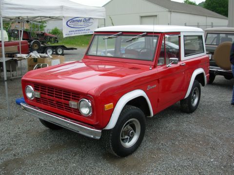 1975 Ford Bronco - A red 1975 Ford Bronco with a white hardtop is seen from a three-quarter front view, parked next to another vehicle.