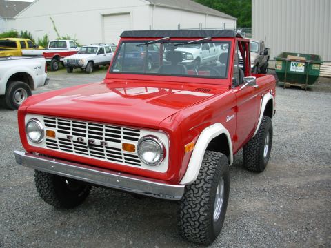 1975 Ford Bronco - A red Ford Bronco is parked in a lot with other vehicles and dumpsters visible. The grille clearly displays the Ford name.