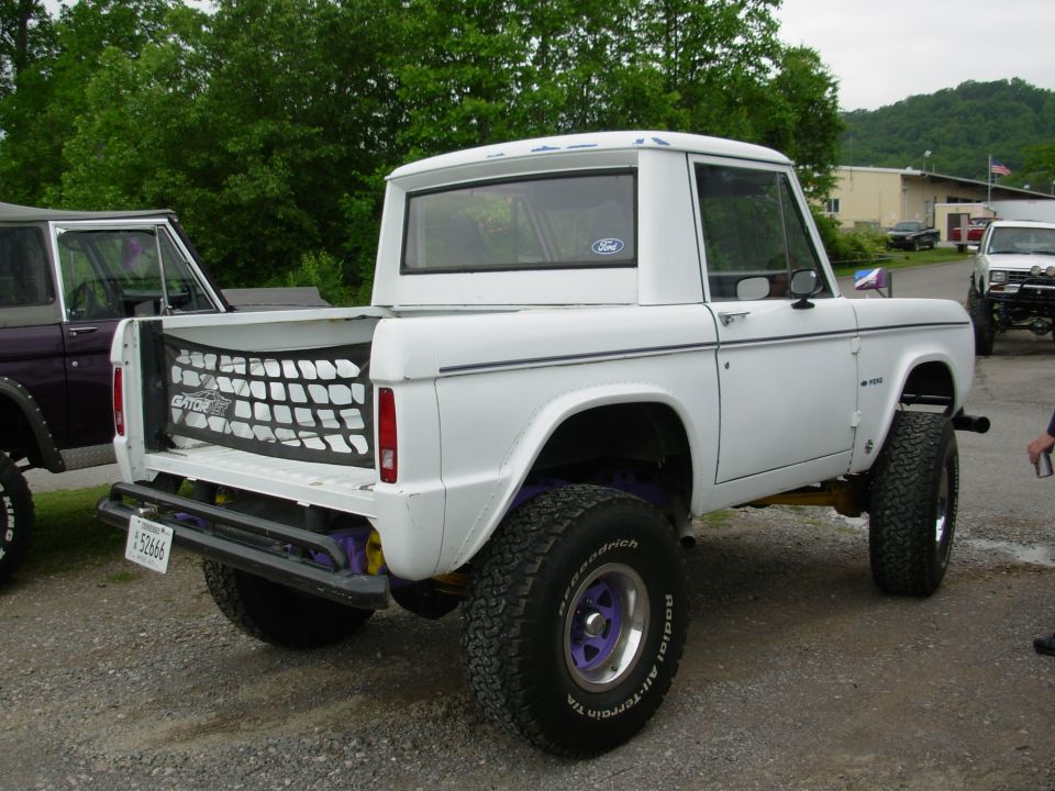 White lifted early Ford Bronco half-cab, tailgate down with mesh net, on gravel lot