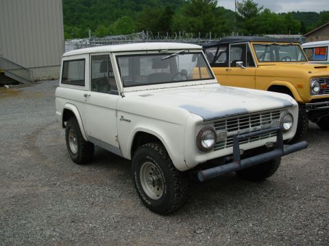 1975 Ford Bronco - A white 1975 Ford Bronco is parked next to a yellow Bronco, with a building and trees in the background.