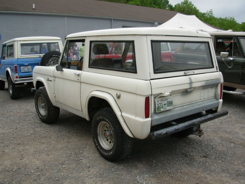 White early Ford Bronco from the rear three-quarter, parked with other classic Broncos on gravel