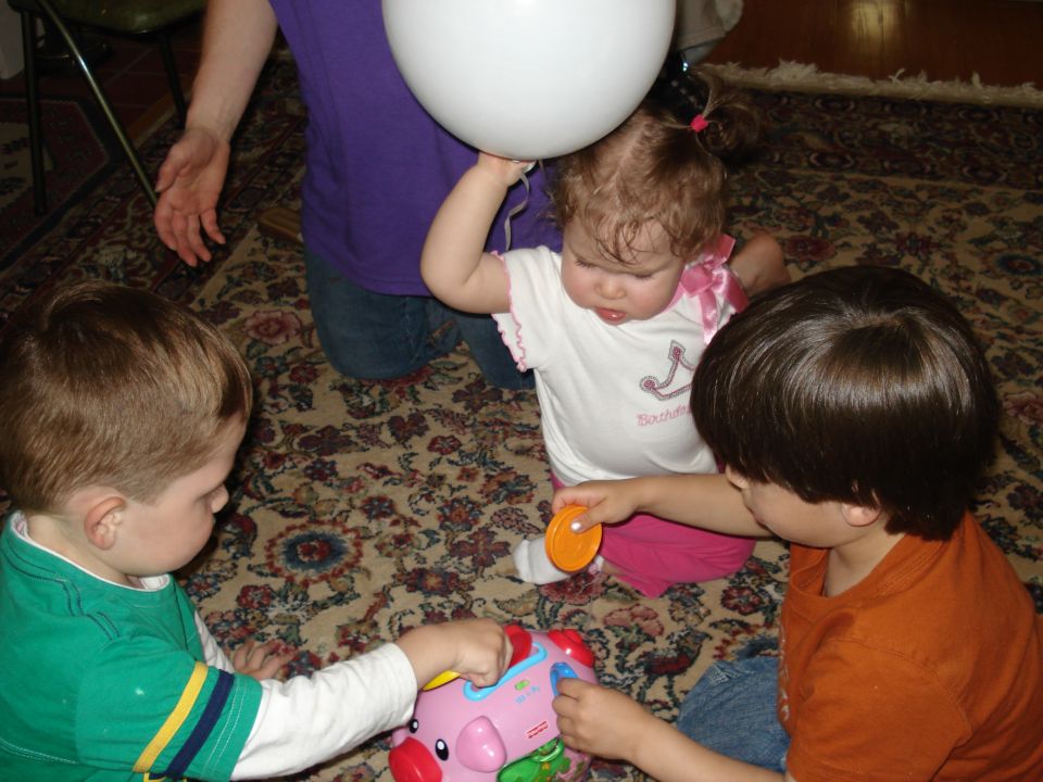 Three young children sit on a patterned rug, playing with a pink toy. One child holds a white balloon
