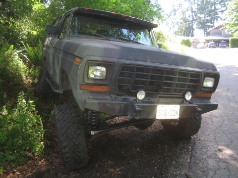 1979 Ford Bronco - A front 3/4 view of a camouflage-painted 1979 Ford Bronco parked on a dirt and asphalt road, surrounded by lush green foliage. The vehicle…
