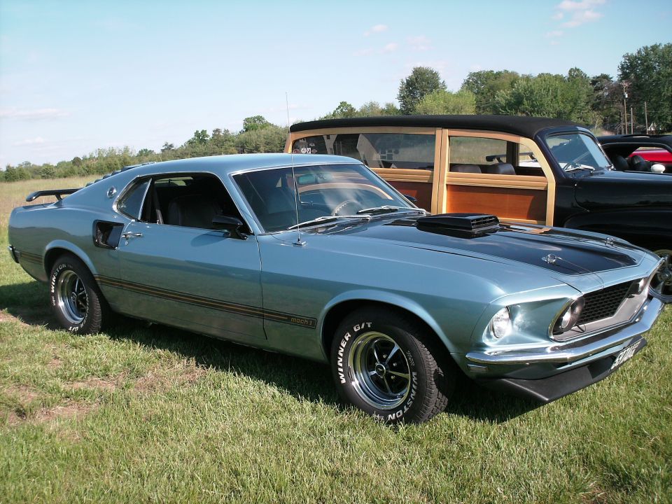 Light blue 1970 Ford Mustang Mach 1 parked on grass, with other vintage cars in the background.