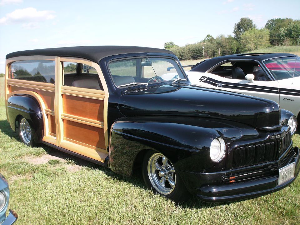 Black 1940s Mercury woody wagon parked on grass next to a 1970 Ford Mustang.