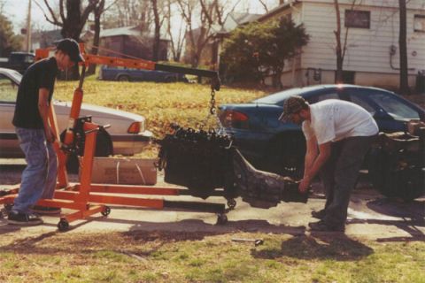 1978 Ford Bronco - An engine is hoisted out of a vehicle, with another car in the background and a person assisting.