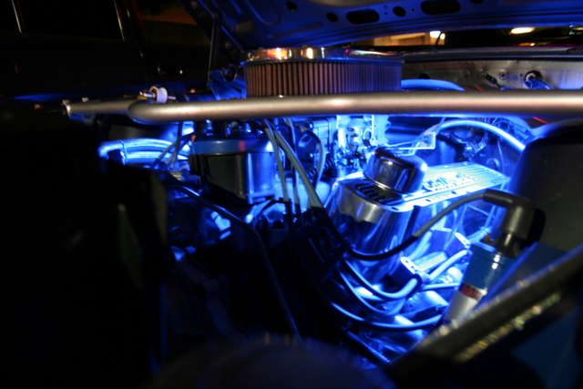 Classic car engine bay with blue LED lighting, showing chrome parts.