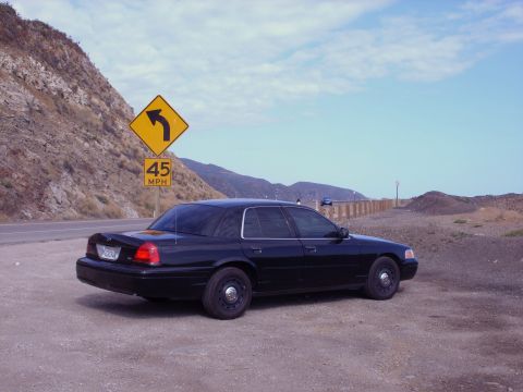 2005 Ford Crown Victoria - A dark sedan is parked on a dirt road with a hill and a yellow road sign in the background.