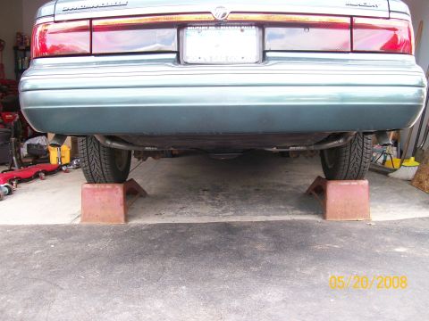 1995 Mercury Grand Marquis - The rear underside of a green sedan is elevated on two red car ramps in a garage. The car's license plate and taillights are visible.