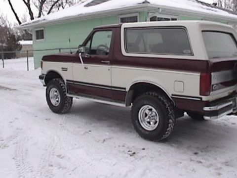 1991 Ford Bronco - A two-tone maroon and cream Ford Bronco is parked on a snow-covered surface in front of a light green house. The vehicle is viewed from the…