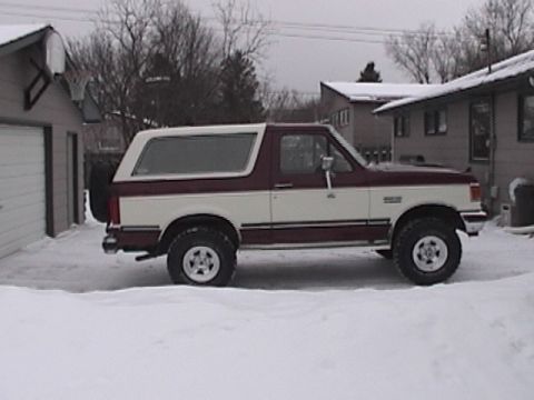 1991 Ford Bronco - A two-tone maroon and white Ford Bronco is parked in a snowy driveway next to a garage. The vehicle is viewed from the side, showing its ti…