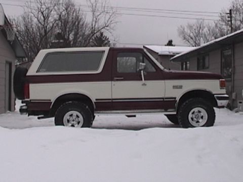 1991 Ford Bronco - A two-tone maroon and white 1991 Ford Bronco is parked in the snow, viewed from the side. The spare tire is mounted on the rear driver's si…