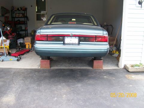 1995 Mercury Grand Marquis - Rear view of a teal/light-green Mercury Grand Marquis LS sedan parked with its rear wheels on wooden ramps in an open garage bay. Full-widt…
