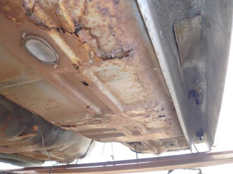 1975 Ford Ranchero - The underside of a vehicle shows significant rust and damage to the floor pan and frame rails. A fuel tank is visible hanging below the rus…