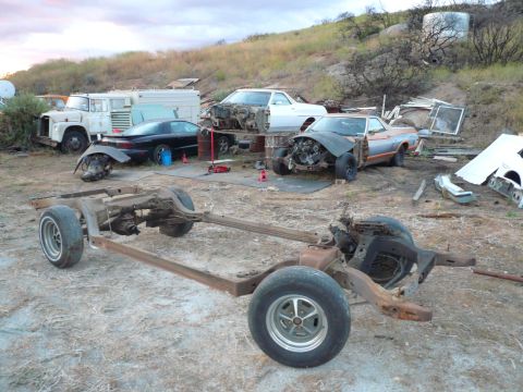 1975 Ford Ranchero - A disassembled vehicle chassis with wheels sits in a junkyard setting, with other car parts and vehicles visible in the background.