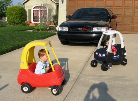 2005 Ford Crown Victoria - Two children's toy cars are parked on a driveway, with a larger black vehicle behind them.