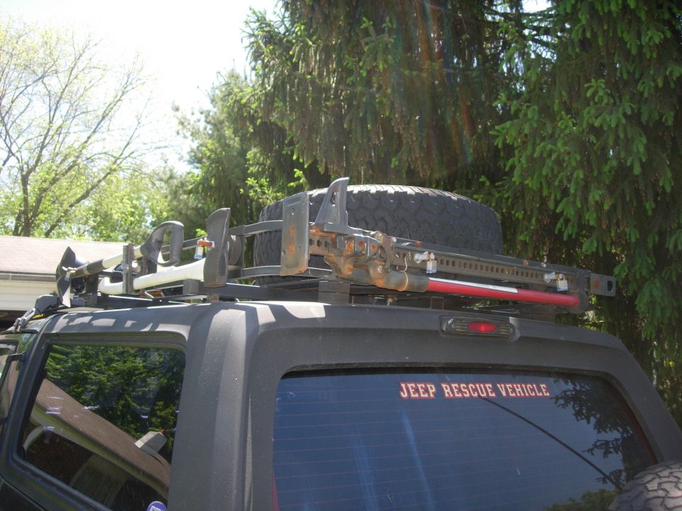 Black SUV with roof rack, spare tire, Hi-Lift jack, axe, shovel, and "JEEP RESCUE VEHICLE" decal.