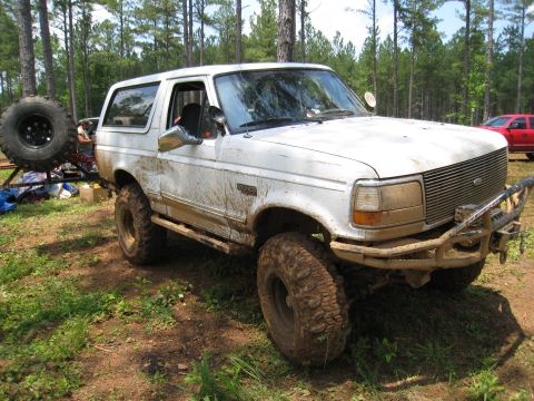 1996 Ford Bronco - A heavily modified 1996 Ford Bronco sits in a muddy, wooded setting, with another red vehicle visible in the background.