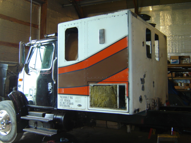 Semi-truck cab with white and orange/brown stripe paint, undergoing apparent repair or modification. Some insulation is visible through an open panel