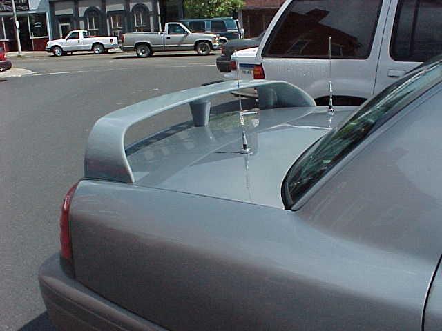 Gray sedan trunk with large aftermarket wing spoiler, cars and storefront behind