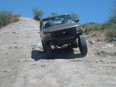 1991 Ford Bronco - A lifted brown Ford Bronco navigates a rocky, off-road trail under a clear blue sky.