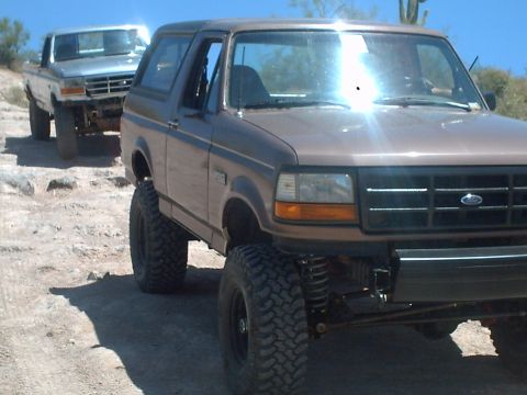 1991 Ford Bronco - Two lifted trucks, a Ford Bronco and an F-Series, are parked on a dirt road under a clear sky.