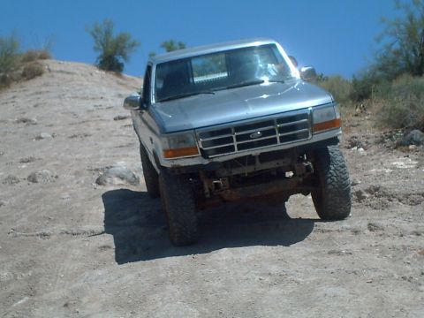 1991 Ford Bronco - A silver pickup truck navigates a rocky, off-road terrain under a clear blue sky.