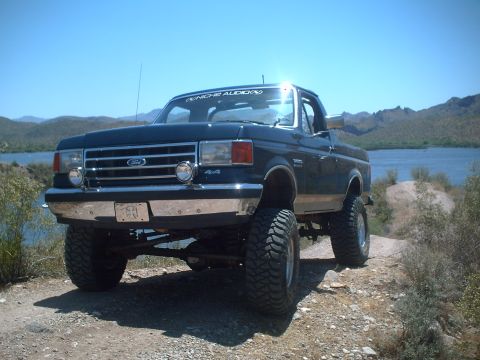 1991 Ford Bronco - Lifted 1991 Ford Bronco with off-road tires sits by a lake under a clear blue sky.