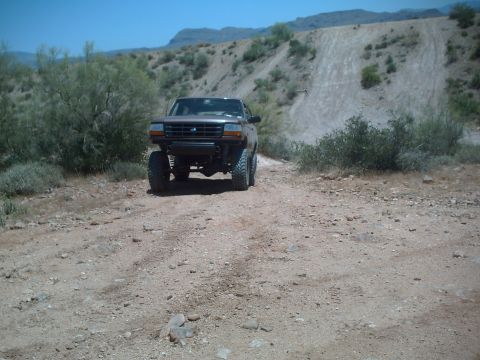 1991 Ford Bronco - A lifted Ford truck drives on a dusty, off-road trail with arid hills in the background.