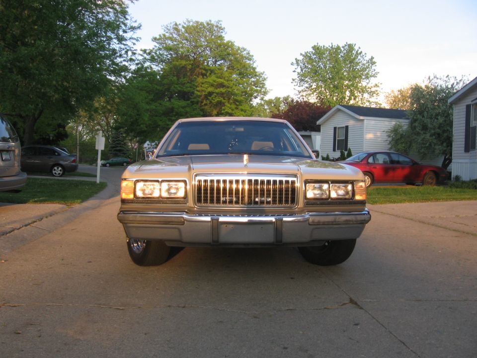 1988 Mercury Grand Marquis front view. Tan paint, chrome grille, quad headlights. Other cars and houses in background