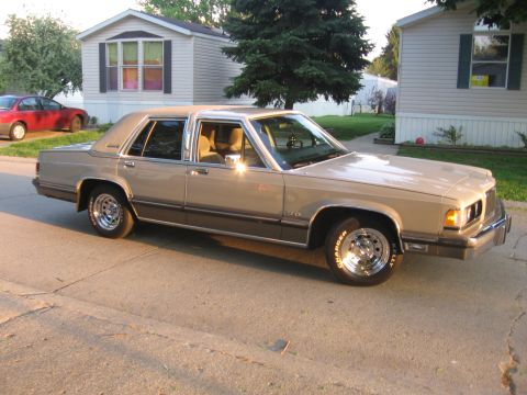 1988 Mercury Grand Marquis - A tan 1988 Mercury Grand Marquis sedan is parked on a street in front of houses, with another red car visible in the background.