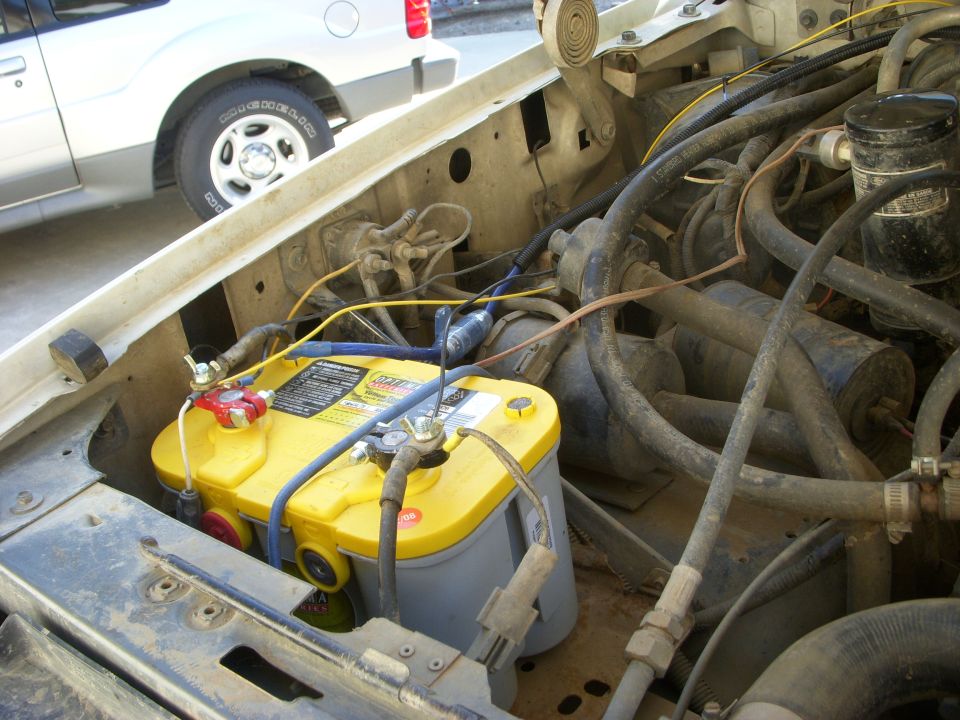 Engine bay with yellow Optima battery and hoses. White SUV in background