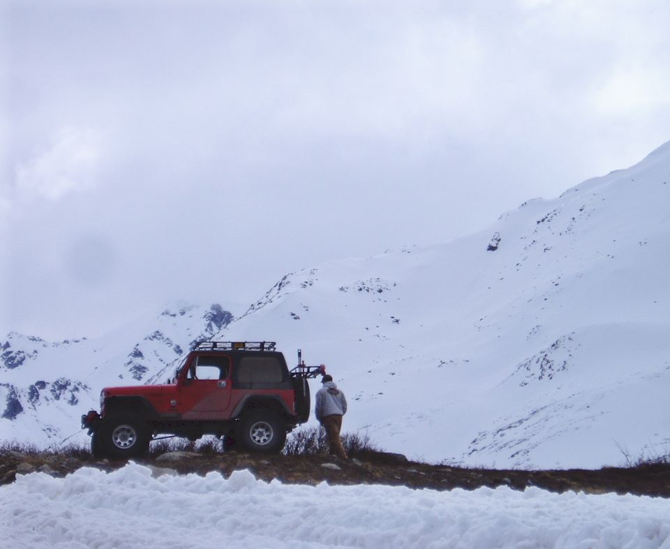 Red Jeep Wrangler parked on a snowy mountain road, side view. A person stands by the rear