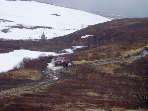 2000 Ford E350 - A red SUV navigates a challenging off-road trail with snow and mud, while another vehicle follows behind.