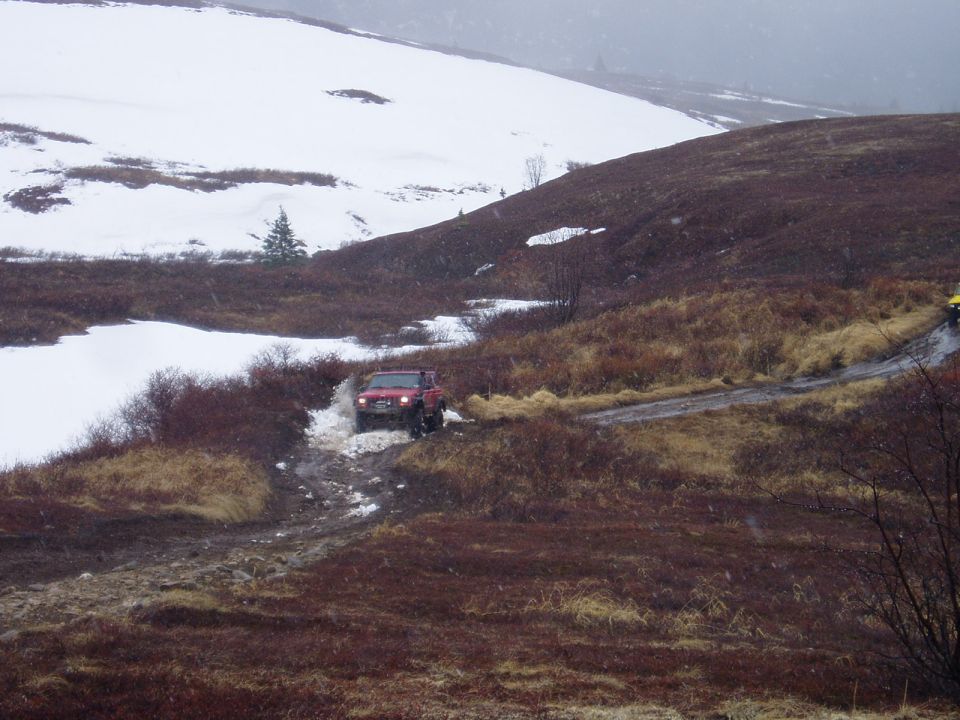 Red SUV driving through mud and snow on a dirt road. Another yellow vehicle is visible further up the road