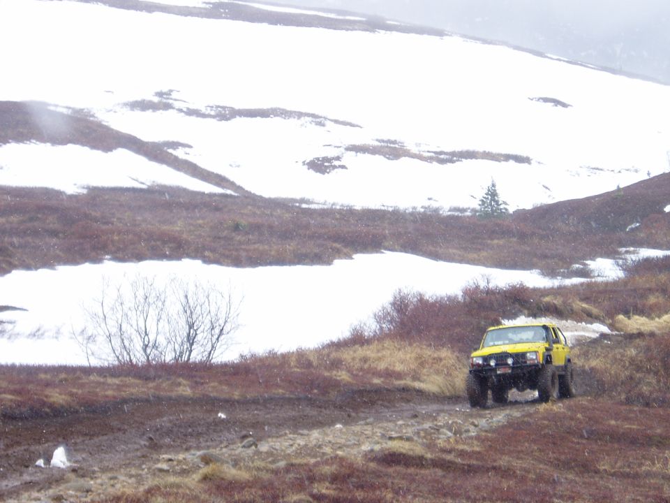 Yellow SUV with large tires on a muddy dirt road with snow-covered hills in the background