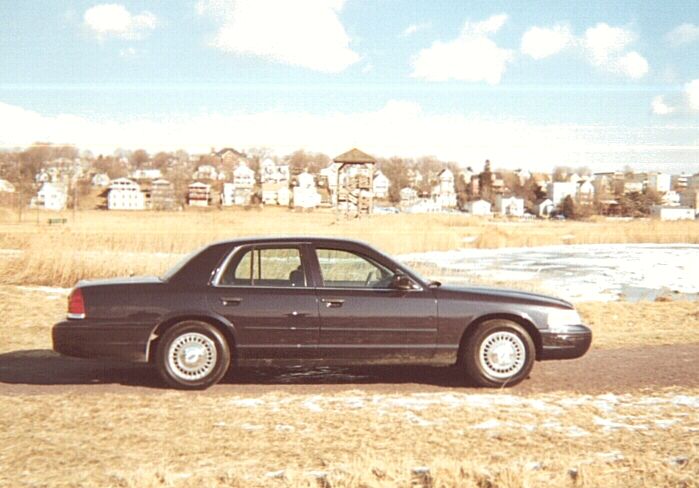 Dark Mercury Grand Marquis sedan in profile parked on dirt by a frozen marsh with houses behind
