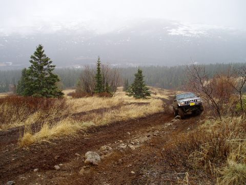 2000 Ford E350 - A Jeep Grand Cherokee tackles a challenging muddy trail in a scenic, overcast landscape.