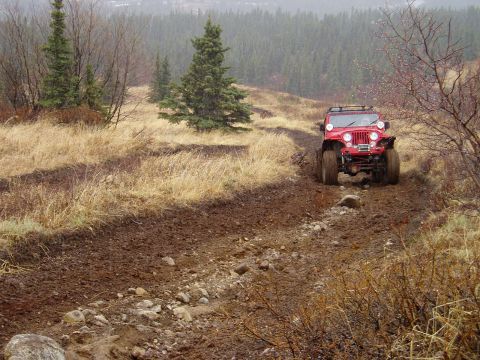 2000 Ford E350 - Red Jeep Wrangler navigates a challenging muddy off-road trail in a wooded area.