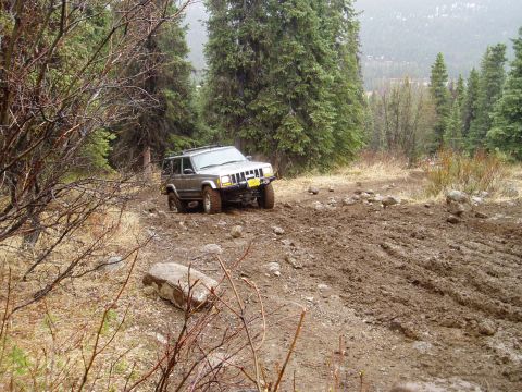 2000 Ford E350 - A Jeep Cherokee XJ navigates a challenging muddy path through a forest.
