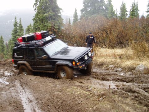 2000 Ford E350 - A black Jeep Cherokee XJ navigates a muddy off-road trail, its tires deep in the mire.