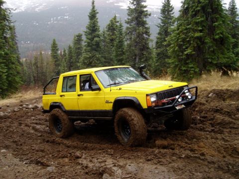 2000 Ford E350 - A yellow Jeep Cherokee XJ navigates a muddy trail in a wooded area.
