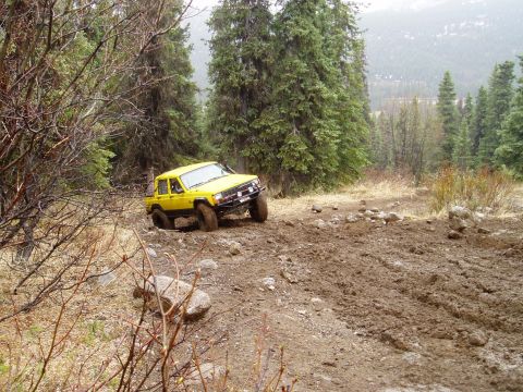 2000 Ford E350 - A yellow off-road vehicle tackles a challenging muddy path through a forest landscape.