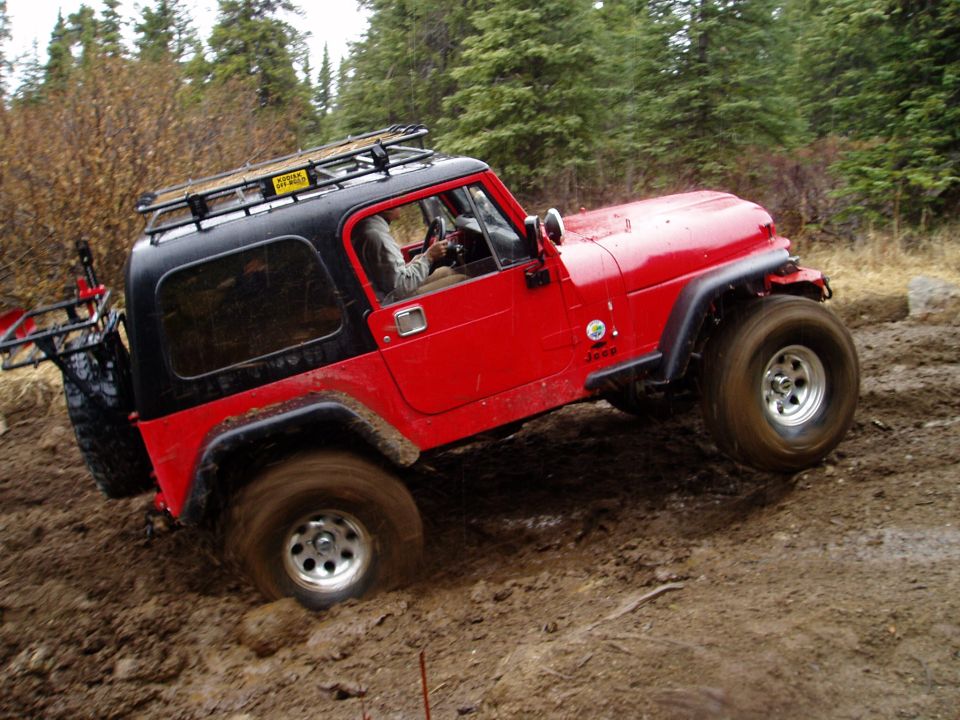 Red Jeep Wrangler with black top and roof rack stuck in mud on a wooded trail