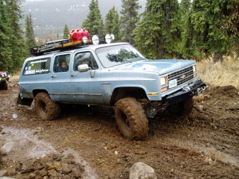 2000 Ford E350 - This modified Chevrolet Suburban is shown off-roading on a wet, muddy track with trees in the background.