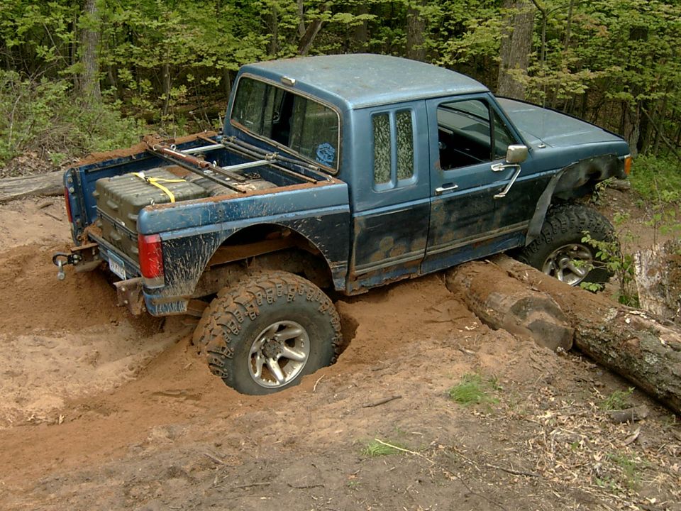 Muddy blue extended-cab off-road pickup climbing over a log on a wooded dirt trail.