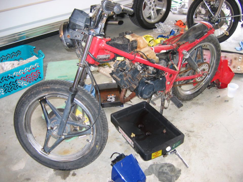 Red motorcycle frame with engine exposed, front wheel and forks visible, oil drain pan on floor