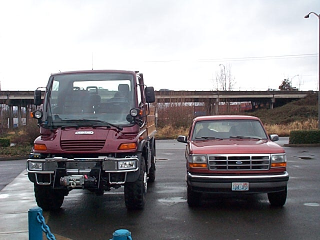 Red Ford Bronco parked next to a taller maroon Unimog truck in a wet parking lot